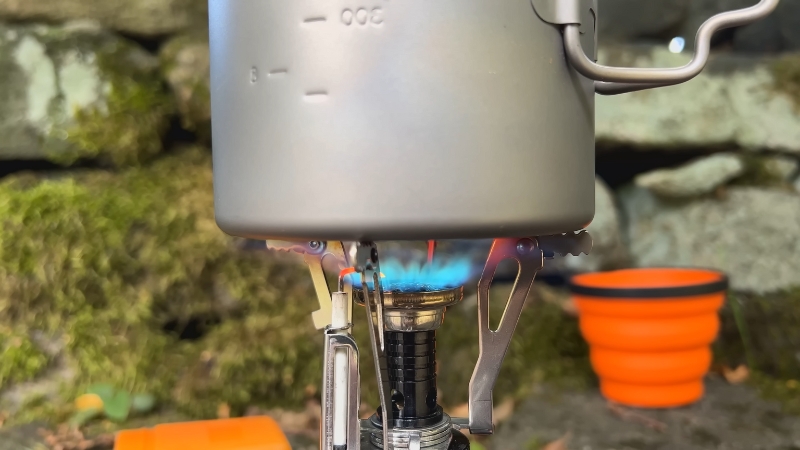 A portable camping stove with a pot, boiling water, set against a natural stone background