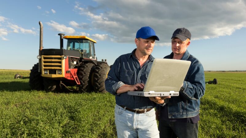 Two farmers check field data on a laptop beside a tractor